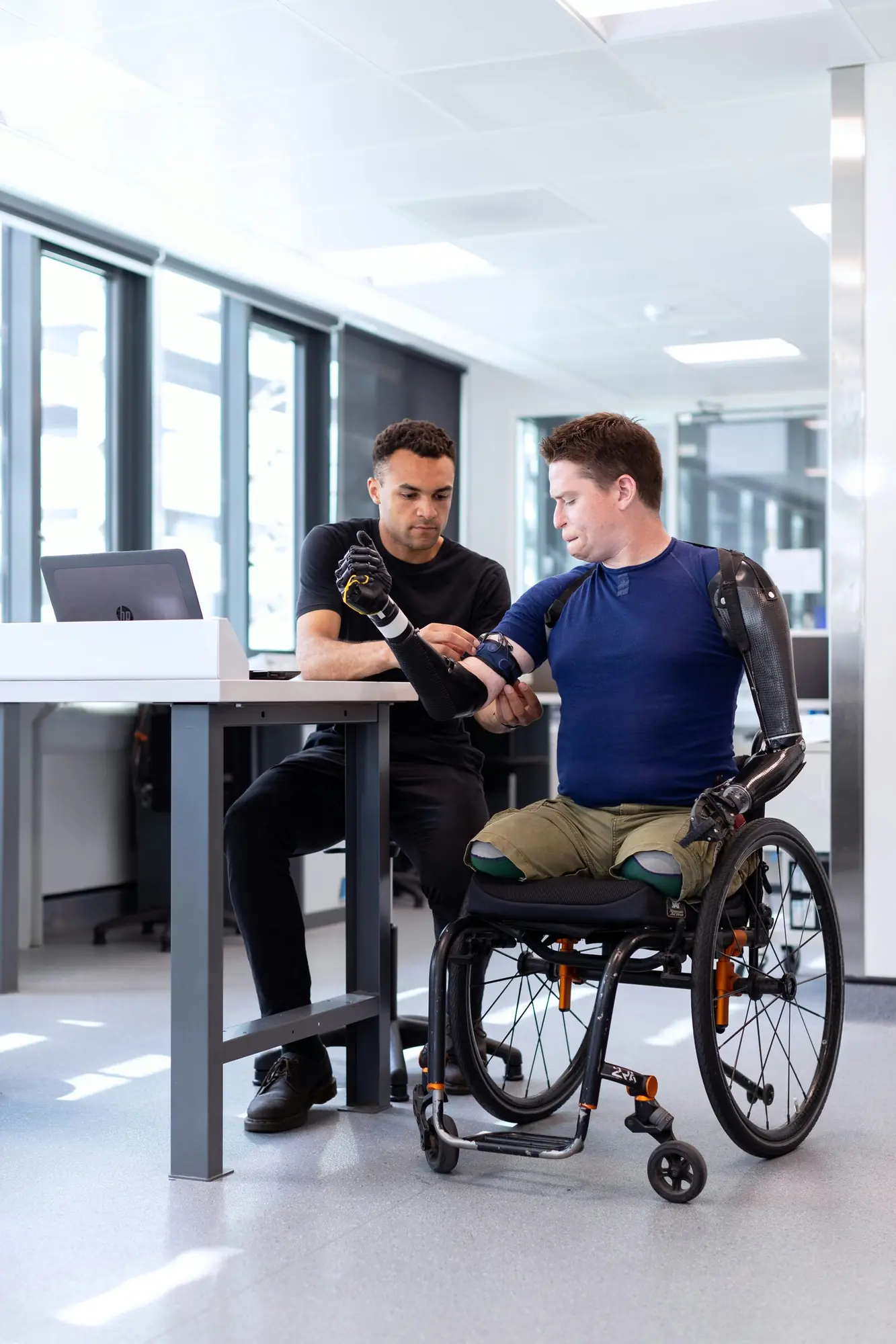 A man using a wheelchair has his prosthetic arm adjusted by a clinician at a desk