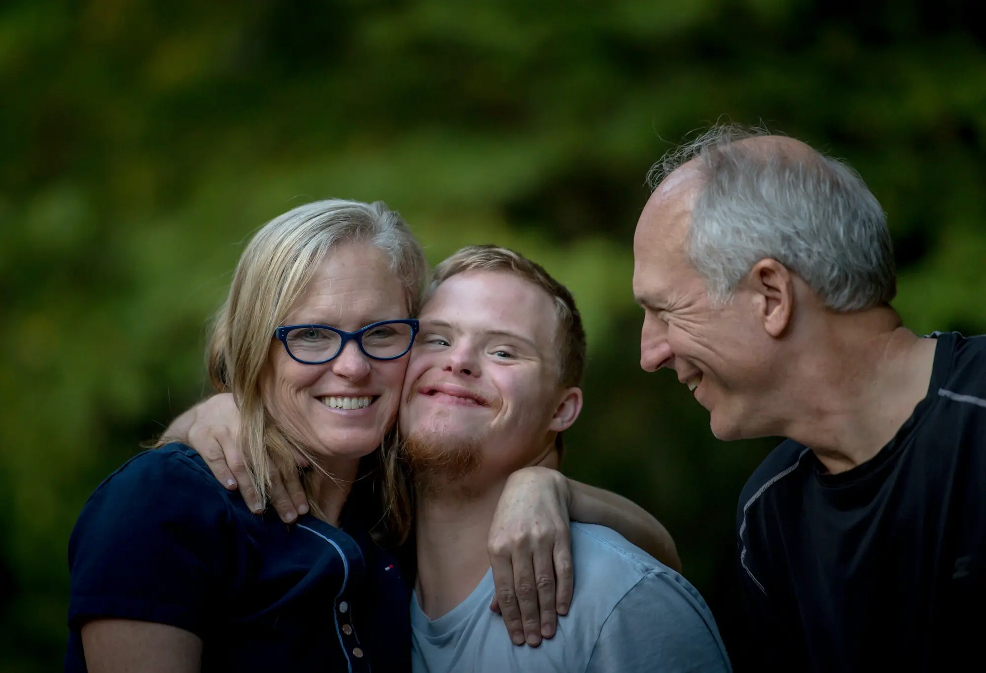 A young man with Down syndrome smiling between two adults who have their arms around him