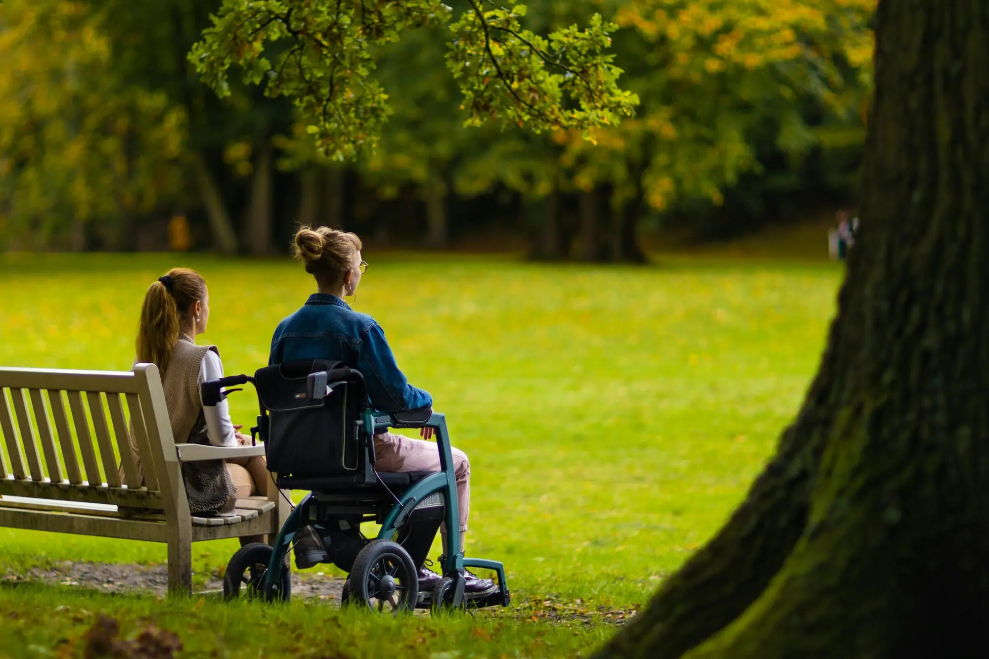 A young woman in a mobility chair sits beside a friend on a park bench in autumn