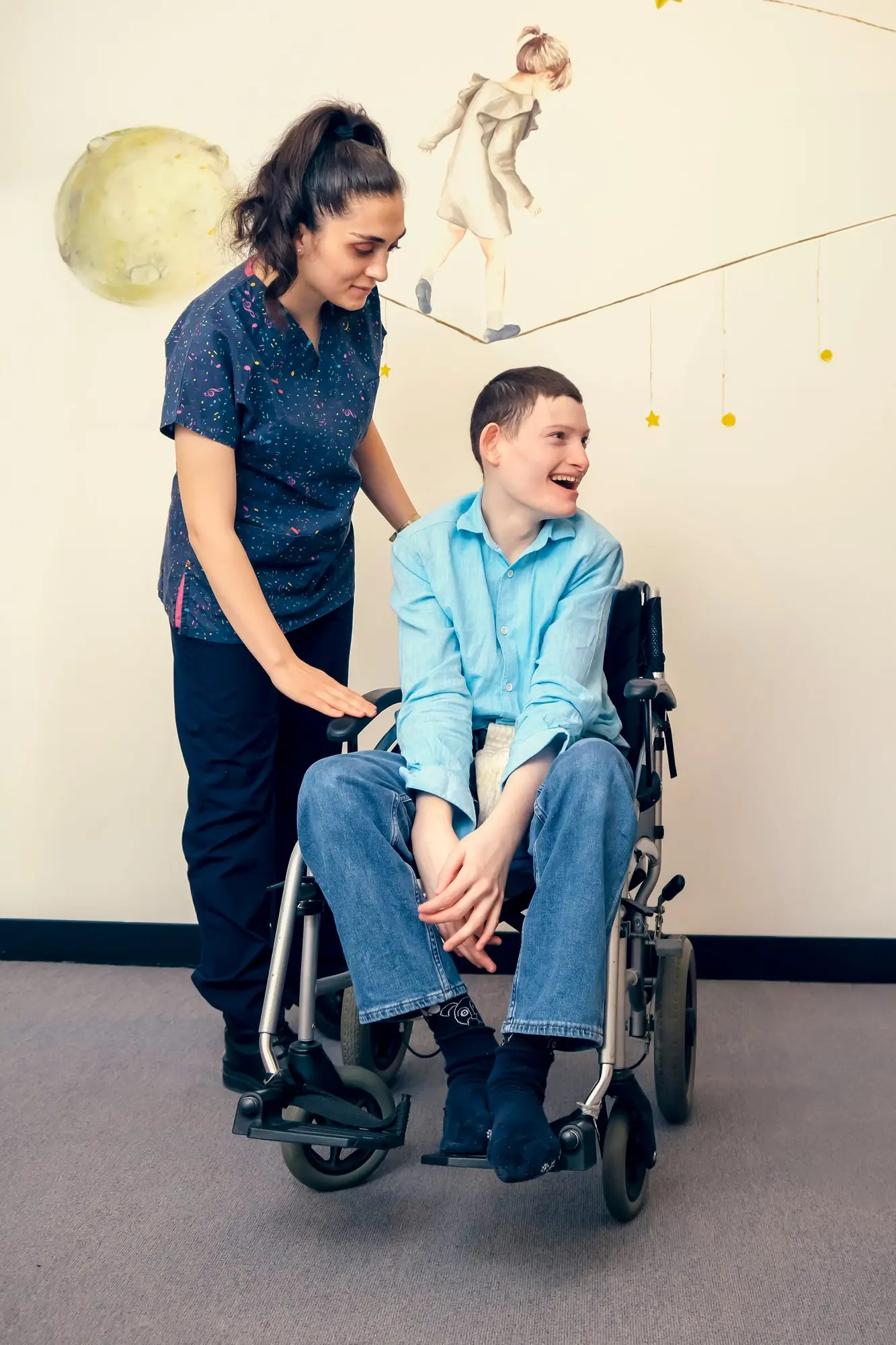 A support worker stands beside a laughing young man in his wheelchair
