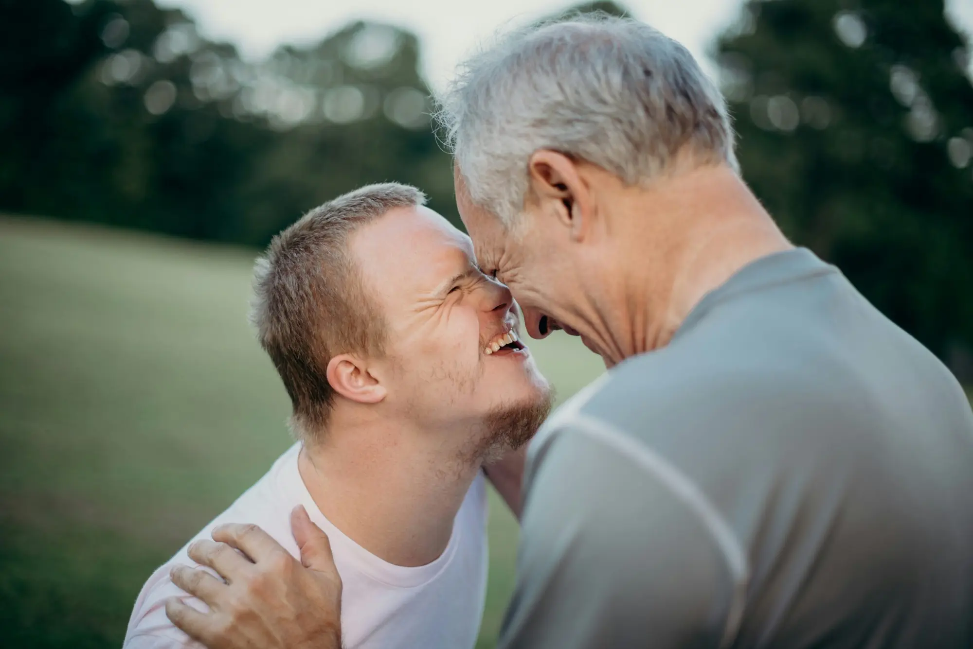 A young man with Down syndrome and an older man laugh together, forehead to forehead, outdoors